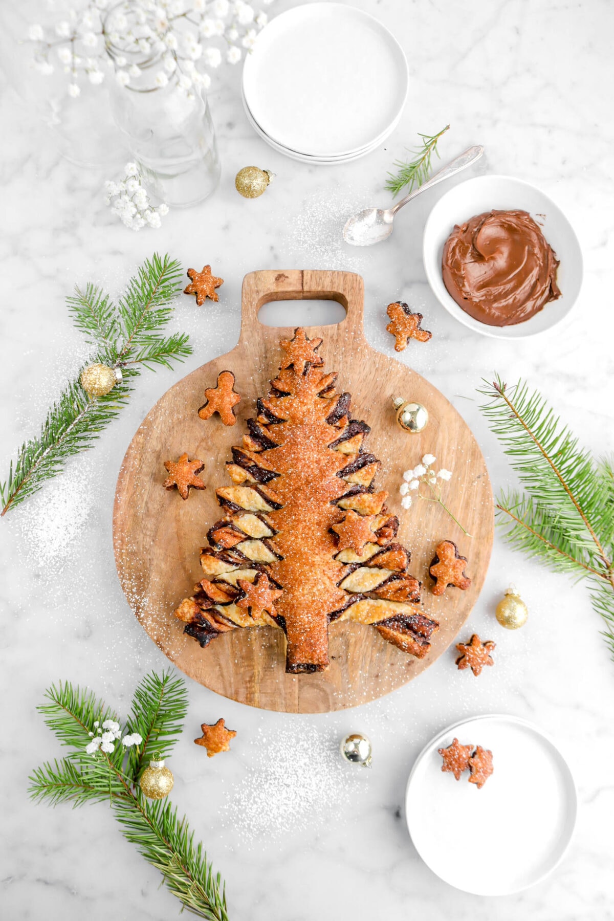 christmas tree tear and share on circle wood board with pine branches, bowl of ganache, gold ornaments, and gingerbread men and snowflake shaped puff pastry bites around.