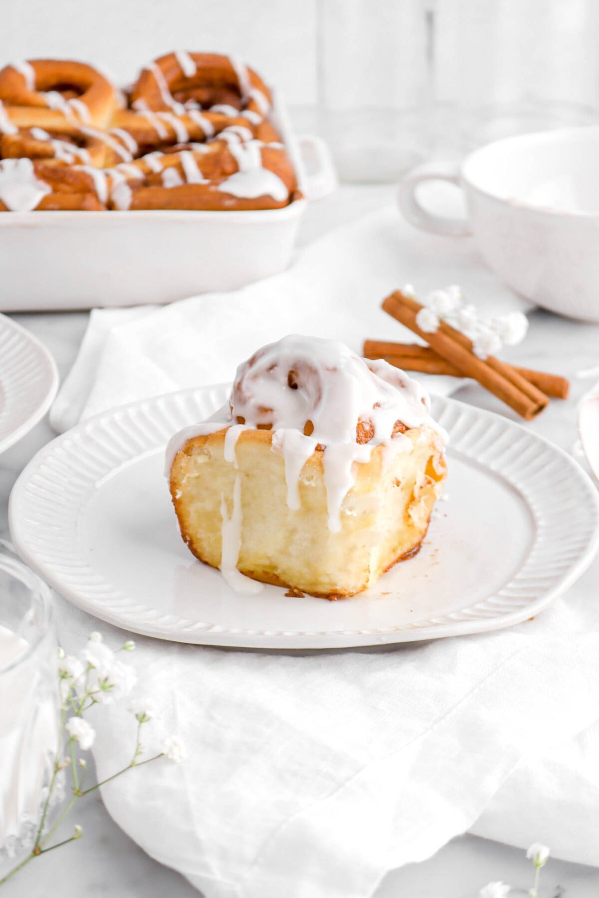 iced cinnamon roll on white plate with two cinnamon sticks behind, a white mug, and casserole of cinnamon rolls.