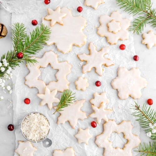 iced snowflake sugar cookies on parchment paper with pine branches, and red cranberries around.