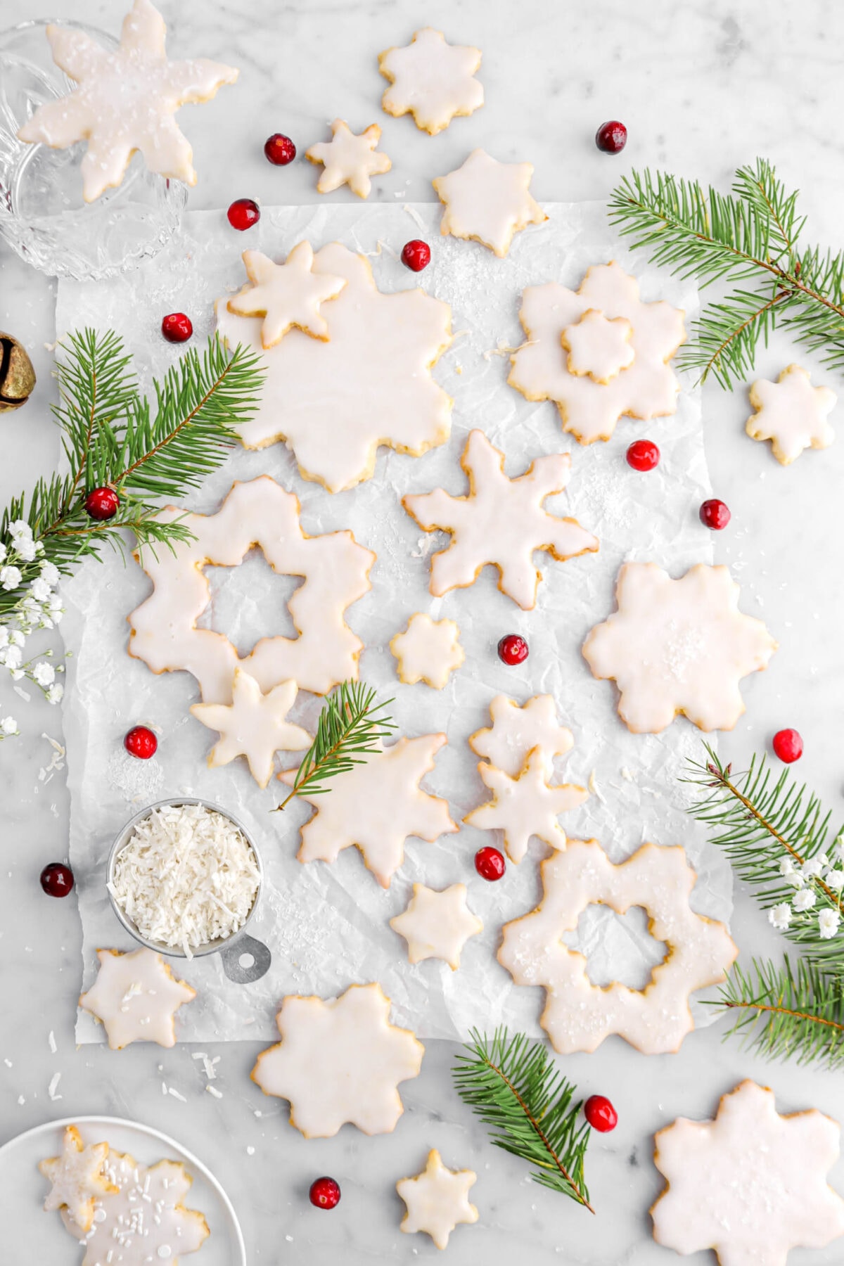 snowflake cookies on parchment paper with greenery around, cranberries, and measuring cup of shredded coconut with a plate of cookies beside.