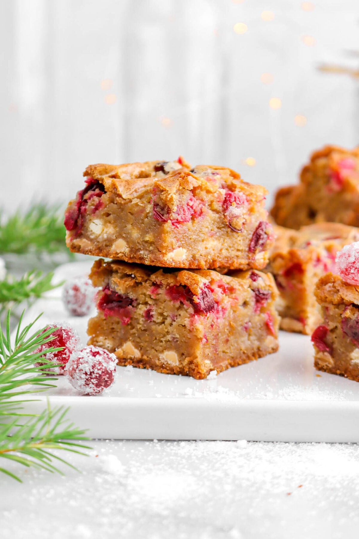 two cranberry white chocolate blondies stacked on white tray with sugared cranberries beside and pine branch.