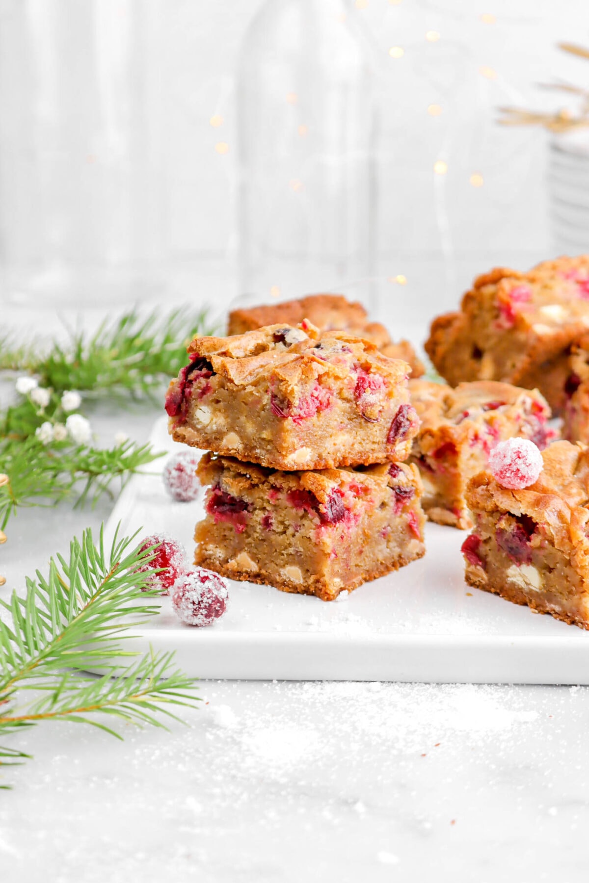 two stacked blondies on white tray with more blondes behind, pine greenery, white flowers, and sugared cranberries beside.
