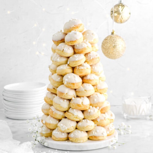 croquembouche on marble cake tray with flowers around, a stack of plates behind, glass of eggnog, and two gold ornaments hanging to the right.