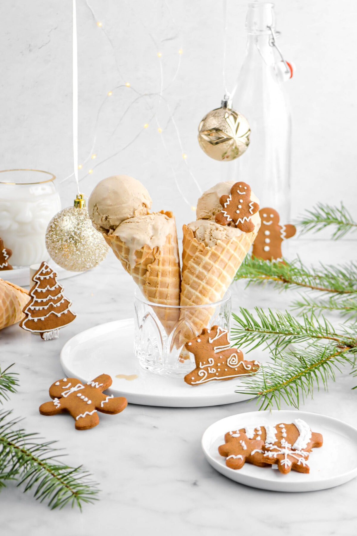 gingerbread ice cream in two waffle cones in a small glass on a white plate with cookies around, pine branches, two gold ornaments, and a glass of milk behind.