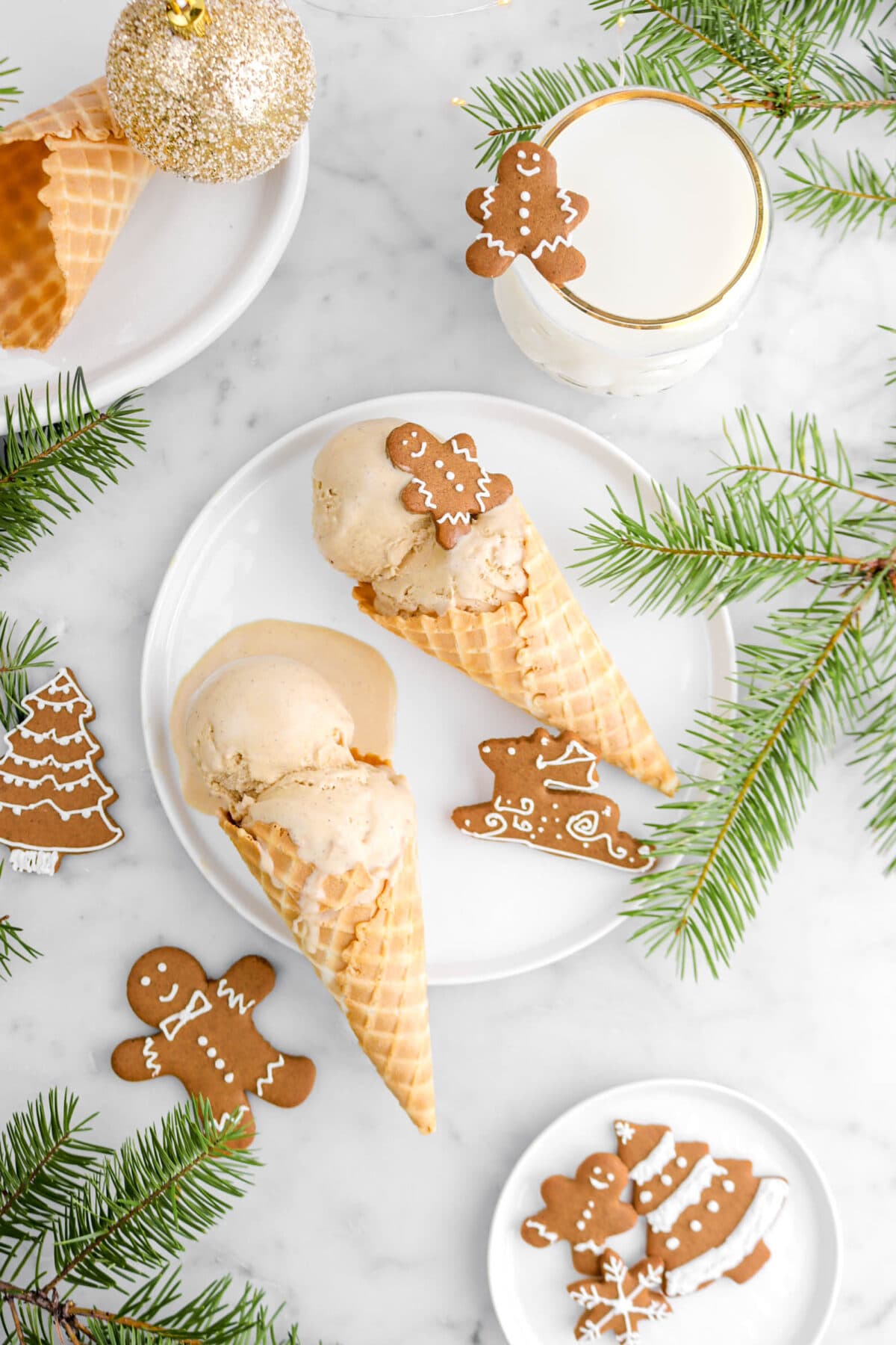 overhead shot of two cones of ice cream on white plate with cookies around, pine branches, and a glass of milk beside.