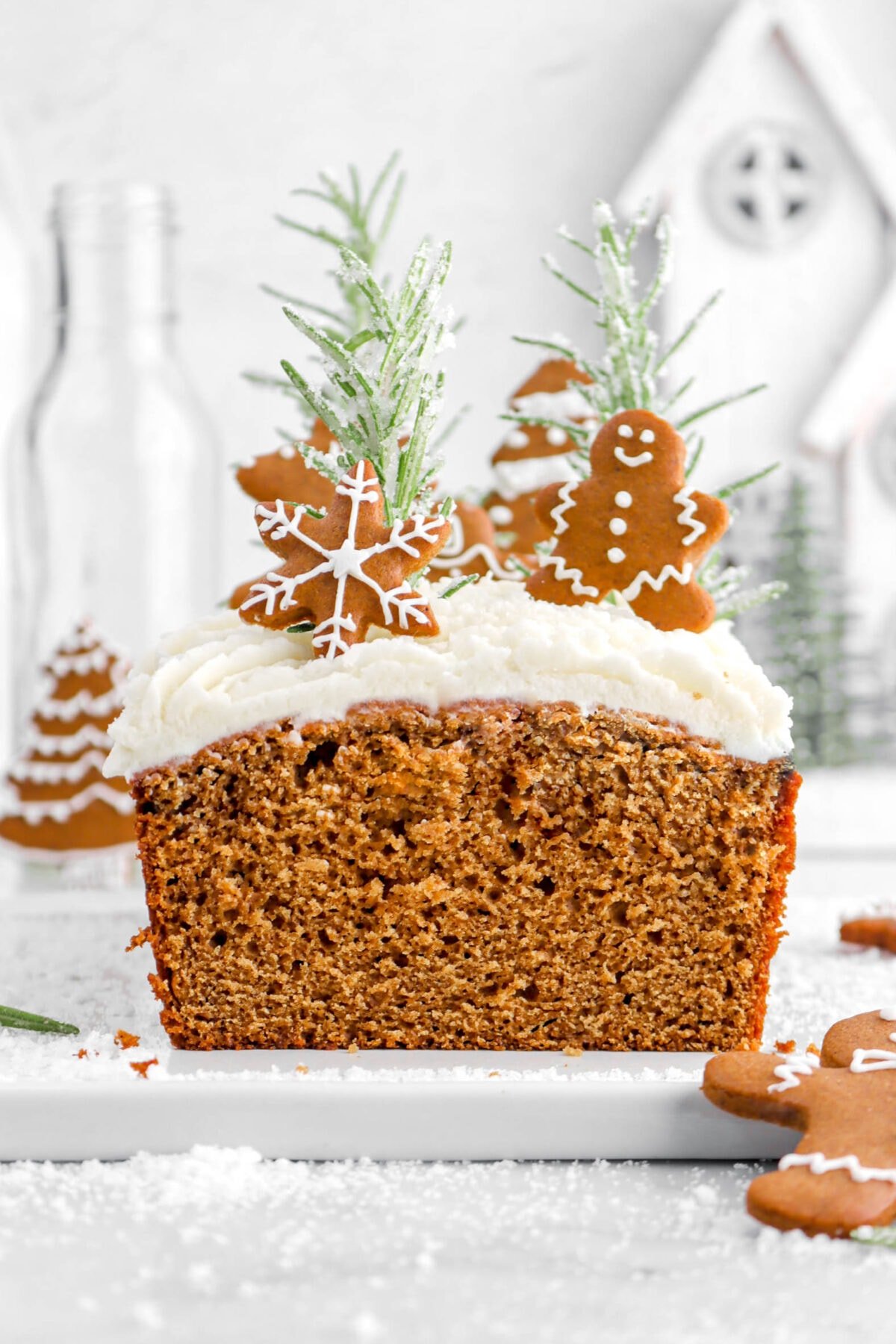 close up of sliced gingerbread loaf cake with slice missing, buttercream on top with decorated mini gingerbread cookies on top, as well as sugared rosemary sprigs.