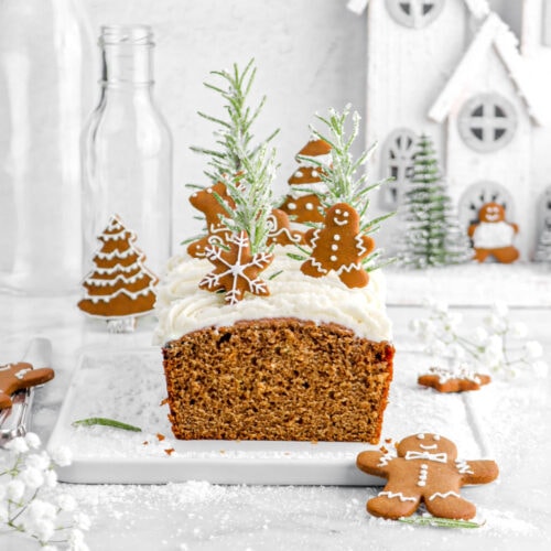 loaf cake on wire serving board with white flowers and sugar around, frosting, mini gingerbread cookies, and sugared rosemary sprigs on top of cake. Christmas village, empty glasses, and more gingerbread cookies behind cake.