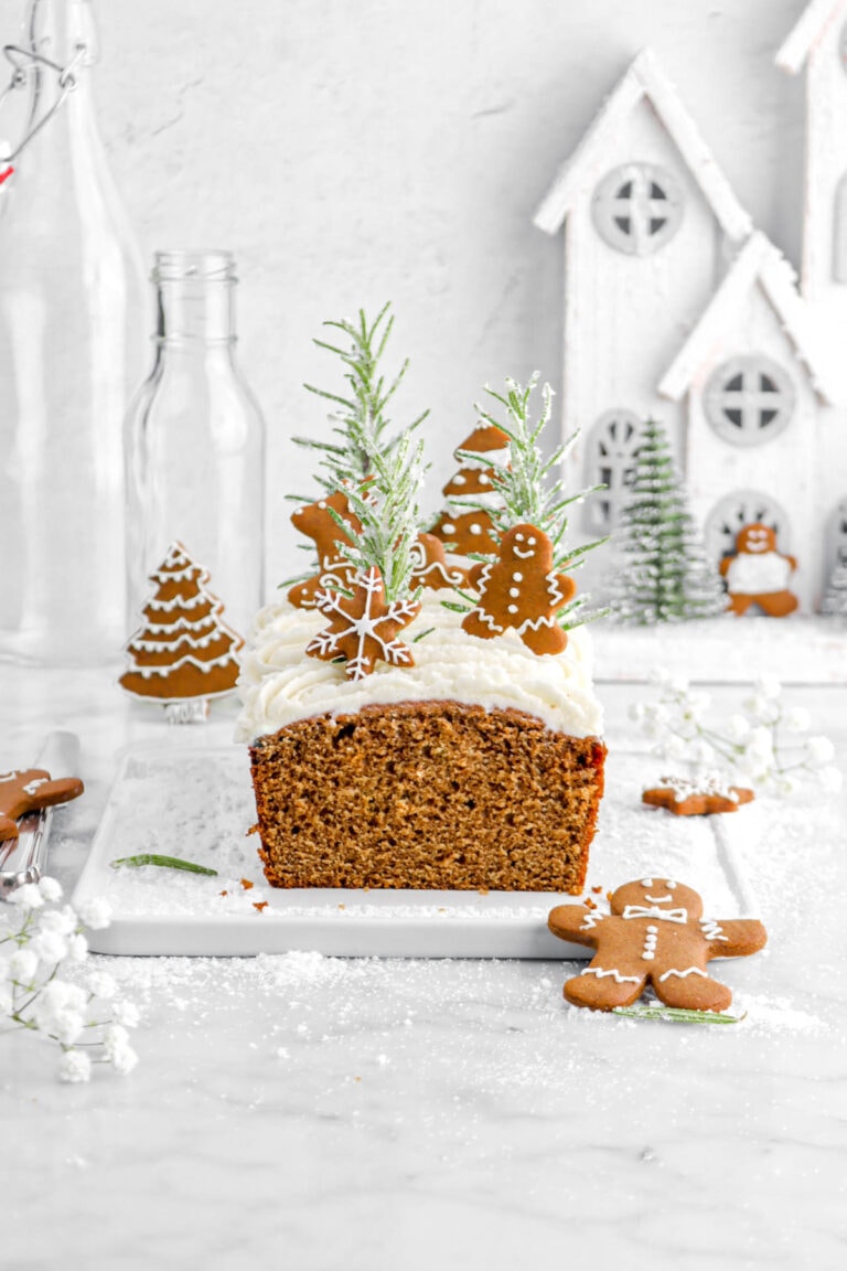 loaf cake on wire serving board with white flowers and sugar around, frosting, mini gingerbread cookies, and sugared rosemary sprigs on top of cake. Christmas village, empty glasses, and more gingerbread cookies behind cake.