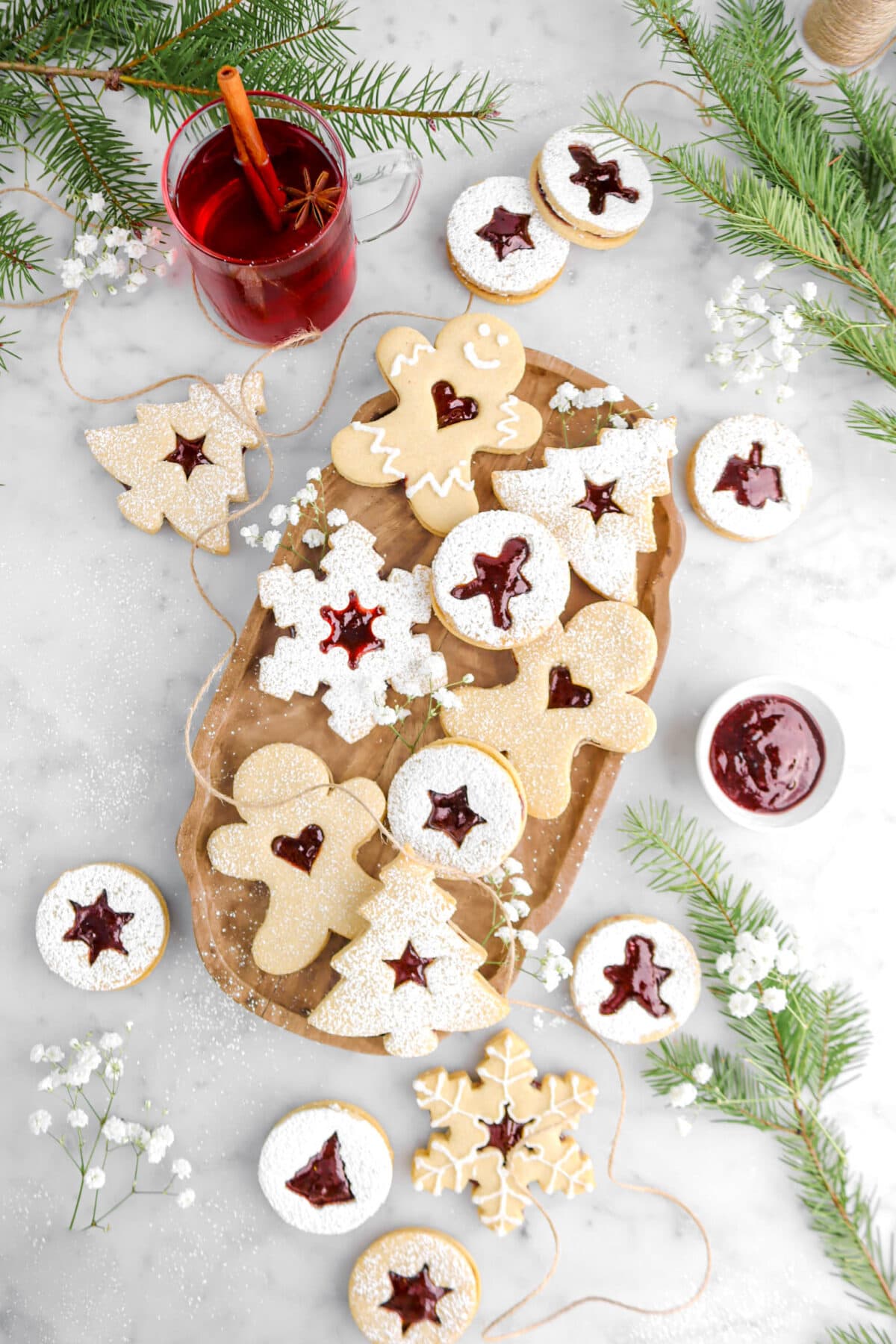 linzer cookies on wood tray with more cookies around, white flowers, pine branches, and glass of mulled wine on marble surface.