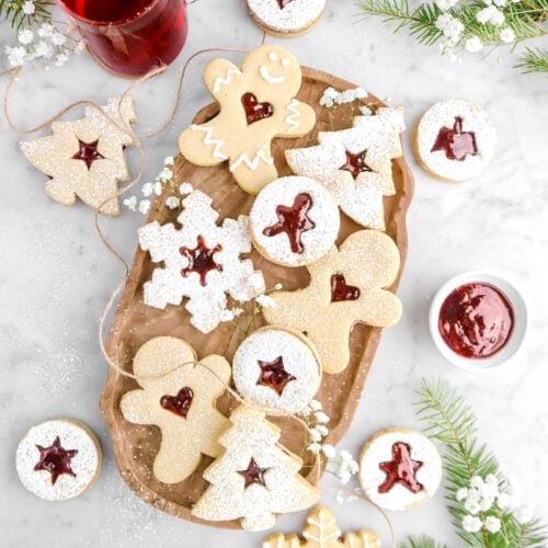 christmas shaped linzer cookies on wood board with ten more around on marble surface, with pine branches, bowl of jelly, and white flowers around.