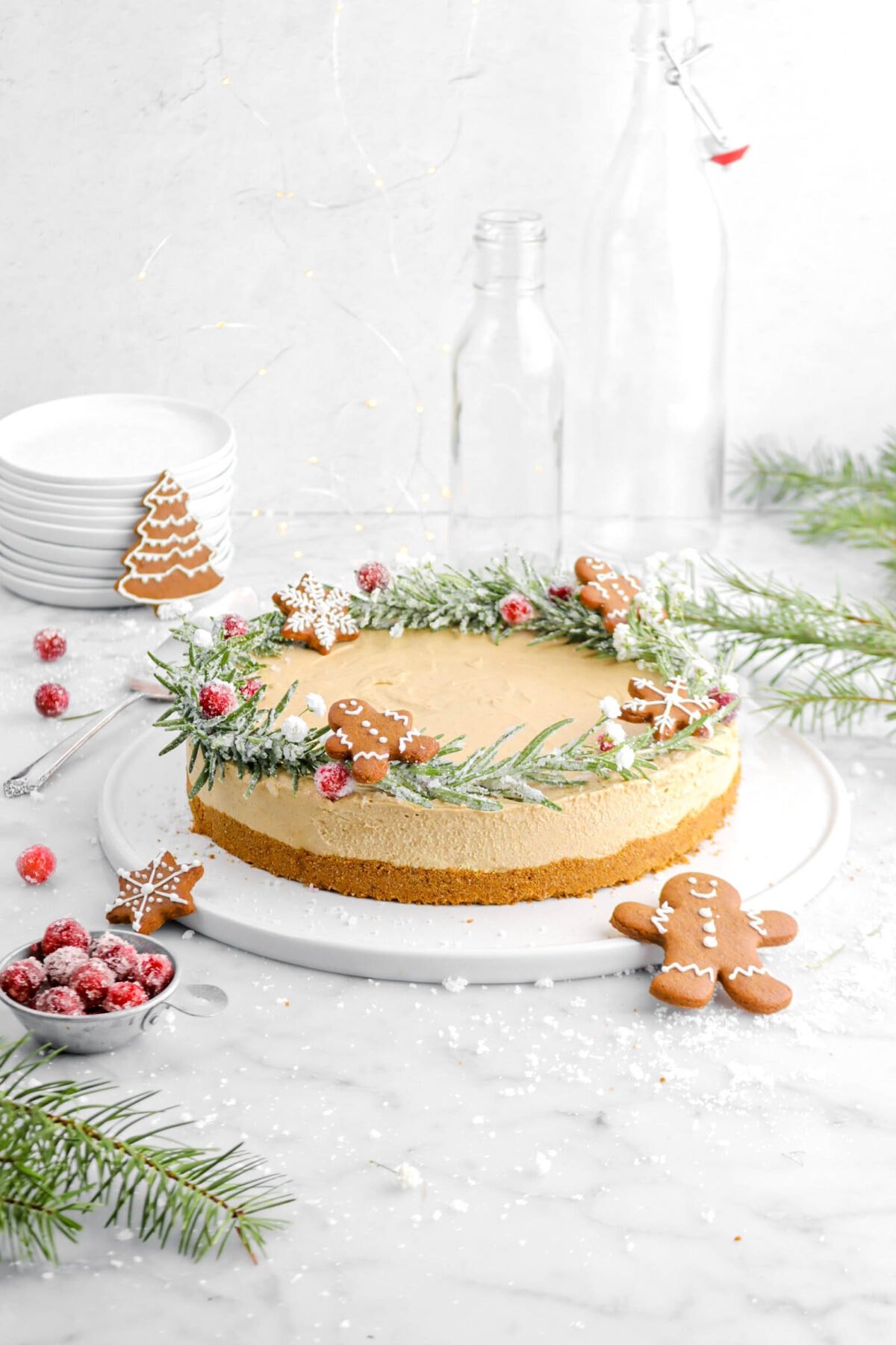 gingerbread cheesecake on round white tray with rosemary crown, sugared cranberries, and mini gingerbread on top with greenery, stack of plates, more sugared cranberries around on marble surface.