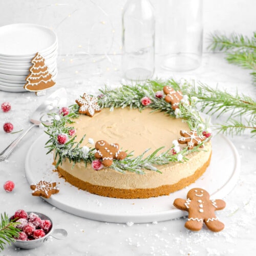 pulled back shot of gingerbread cheesecake on white tray with sugared cranberries, mini gingerbread cookies, and cake knife behind.