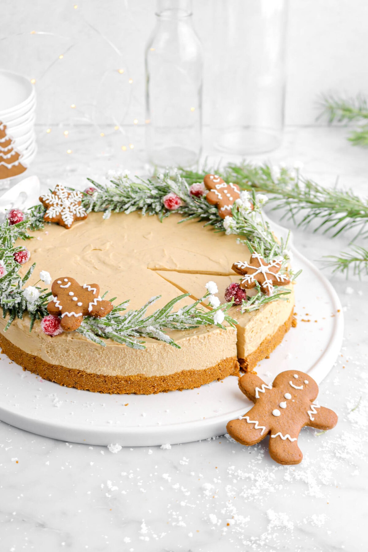 slice cut into cheesecake on white tray with gingerbread cookies on top, sugared rosemary, and sugared cranberries.