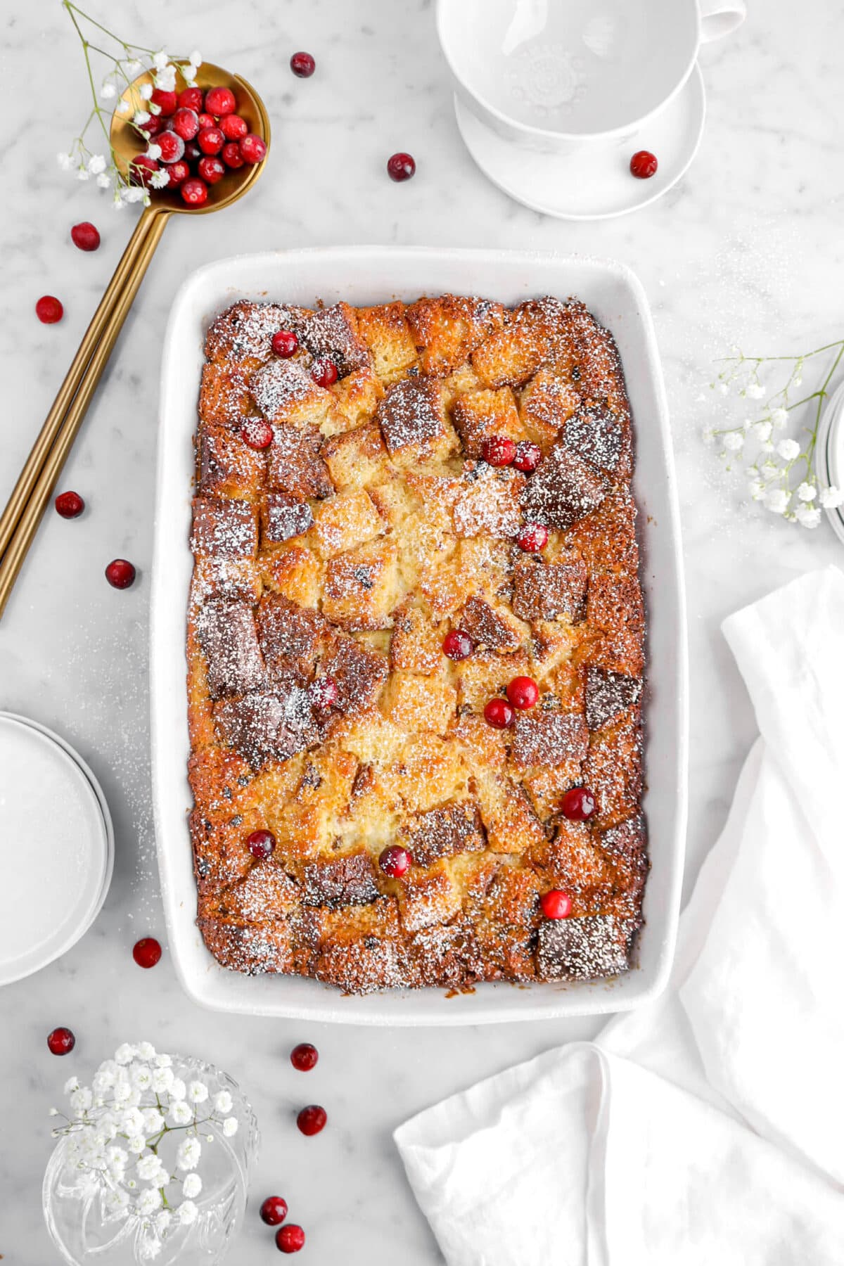 bread pudding with fresh cranberries and powdered sugar on top, with white napkin beside, flowers, and gold serving spoon full of cranberries on marble surface.