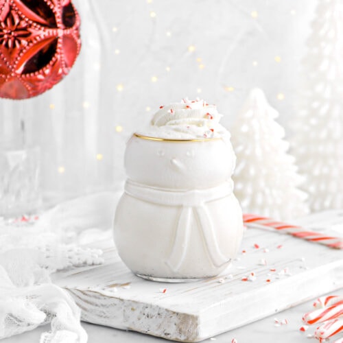 vanilla milkshake in snowman shaped glass on white wood board with whipped cream and crushed peppermint on top, candy canes and white cheesecloth beside, string lights, two white christmas trees, and red ornament behind.