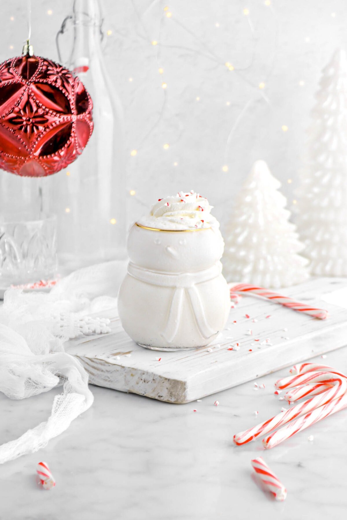 milkshake in snowman shaped glass on a wood board with candy canes and crushed peppermint around, a white cheesecloth beside, a large red ornament hanging behind with two white christmas trees.
