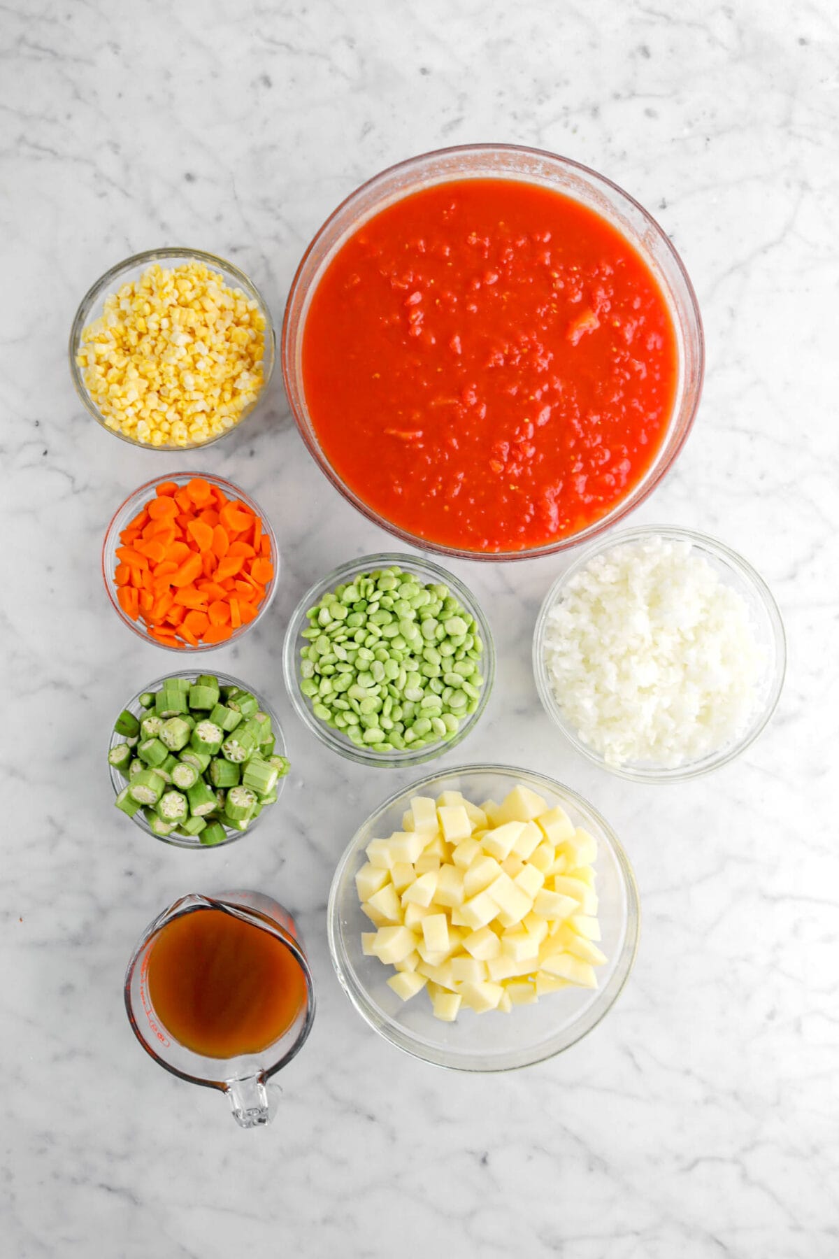 corn, chopped tomatoes, carrots, lima beans, okra, chopped onion, chopped potatoes, and vegetable broth on marble surface.