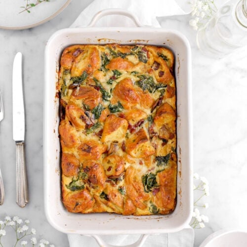 overhead shot of savory breakfast casserole on white napkin with flowers, a plate, and a knife around on marble surface.