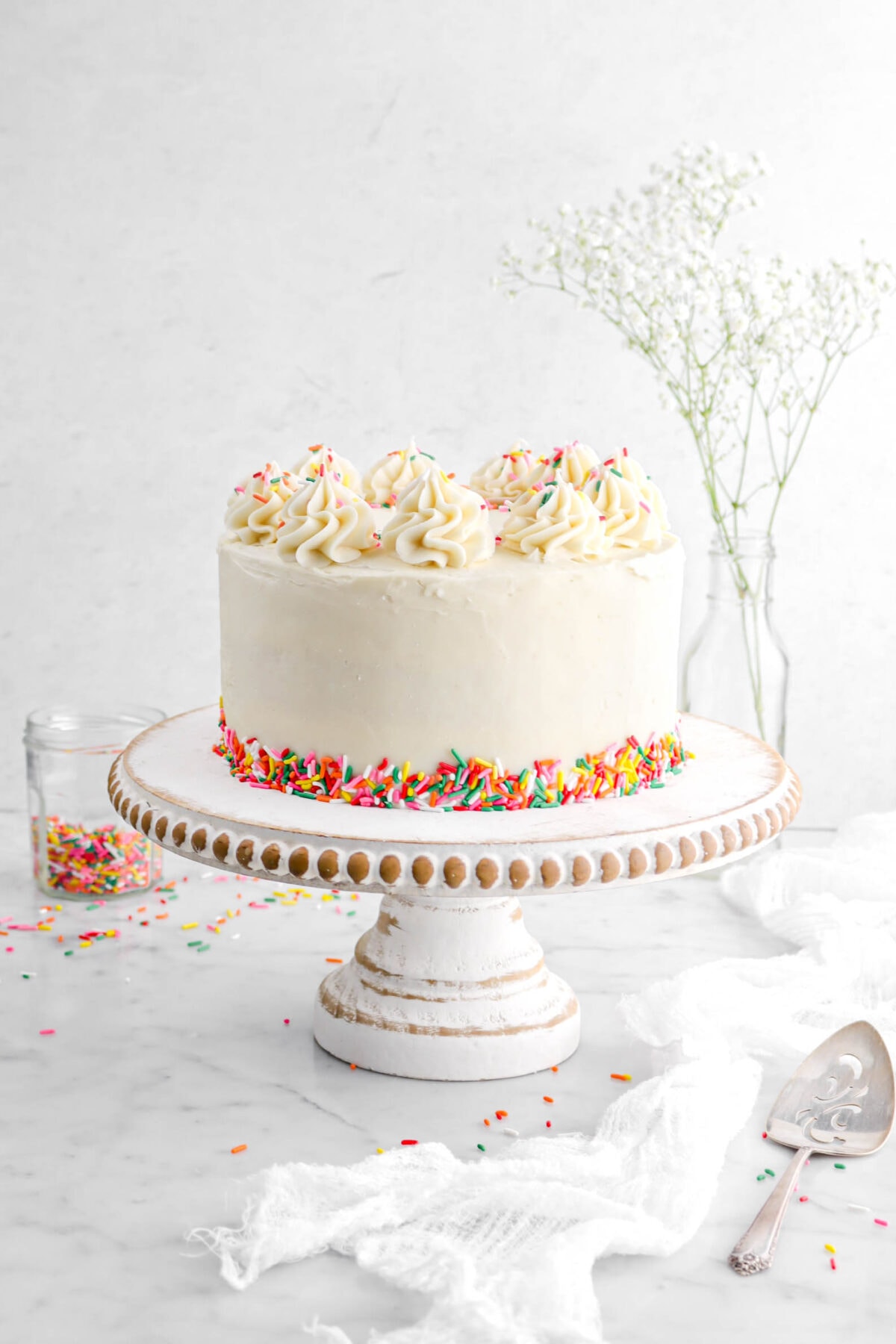 funfetti cake on white washed wood cake stand with frosting piped on the top and sprinkles on the bottom. A white cheesecloth with cake knife beside, jar of rainbow sprinkles and glass f white flowers behind.