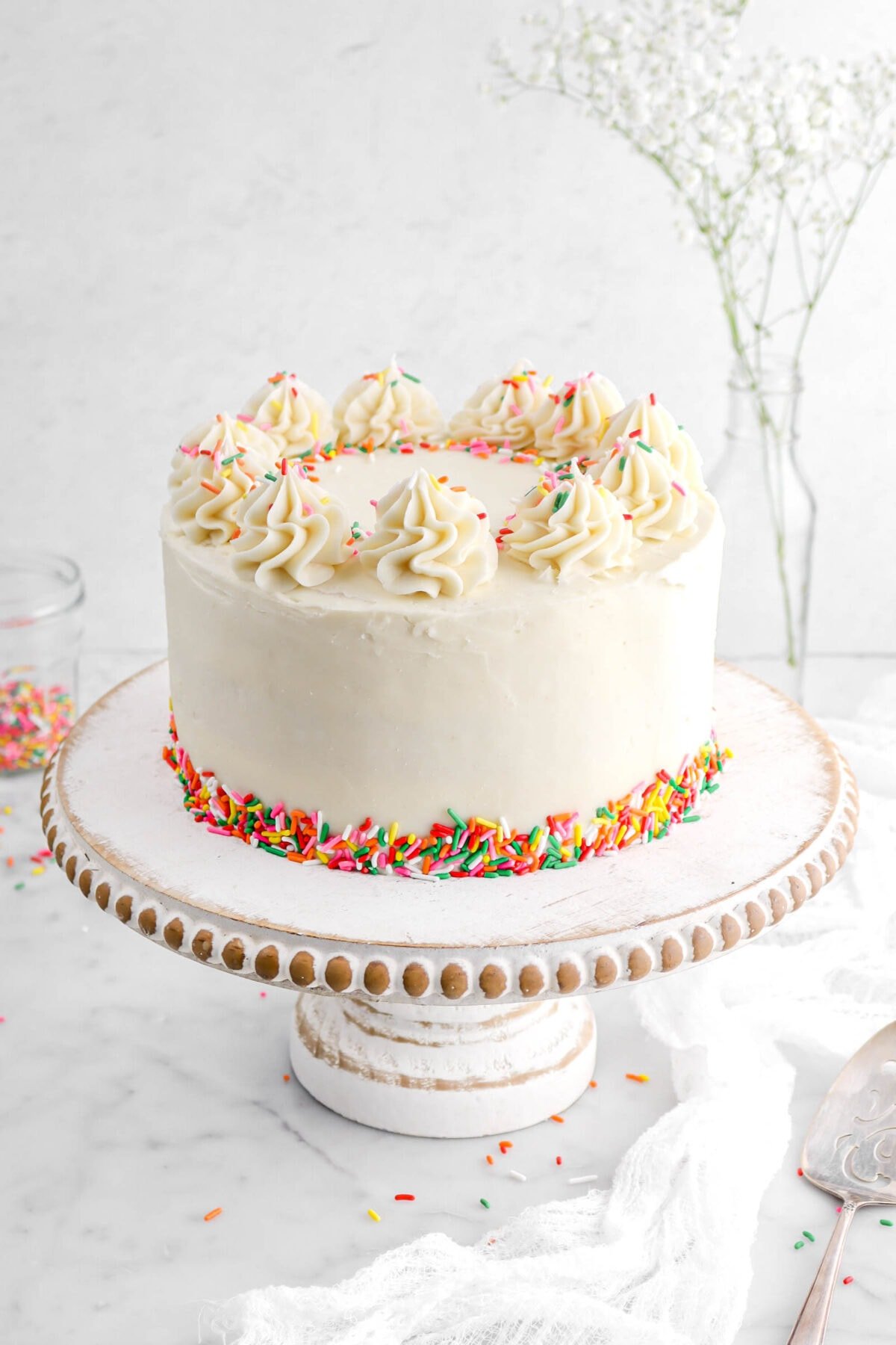 angled close up of funfetti cake on cake stand with rainbow sprinkles around the base of the cake and piped frosting on the top of the cake with extra sprinkles.
