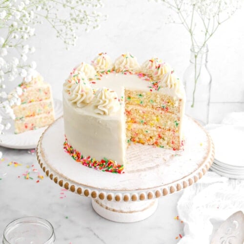 funfetti cake with slices missing on cake sand with flowers around, a slice of cake behind, and a jar of sprinkles in front.