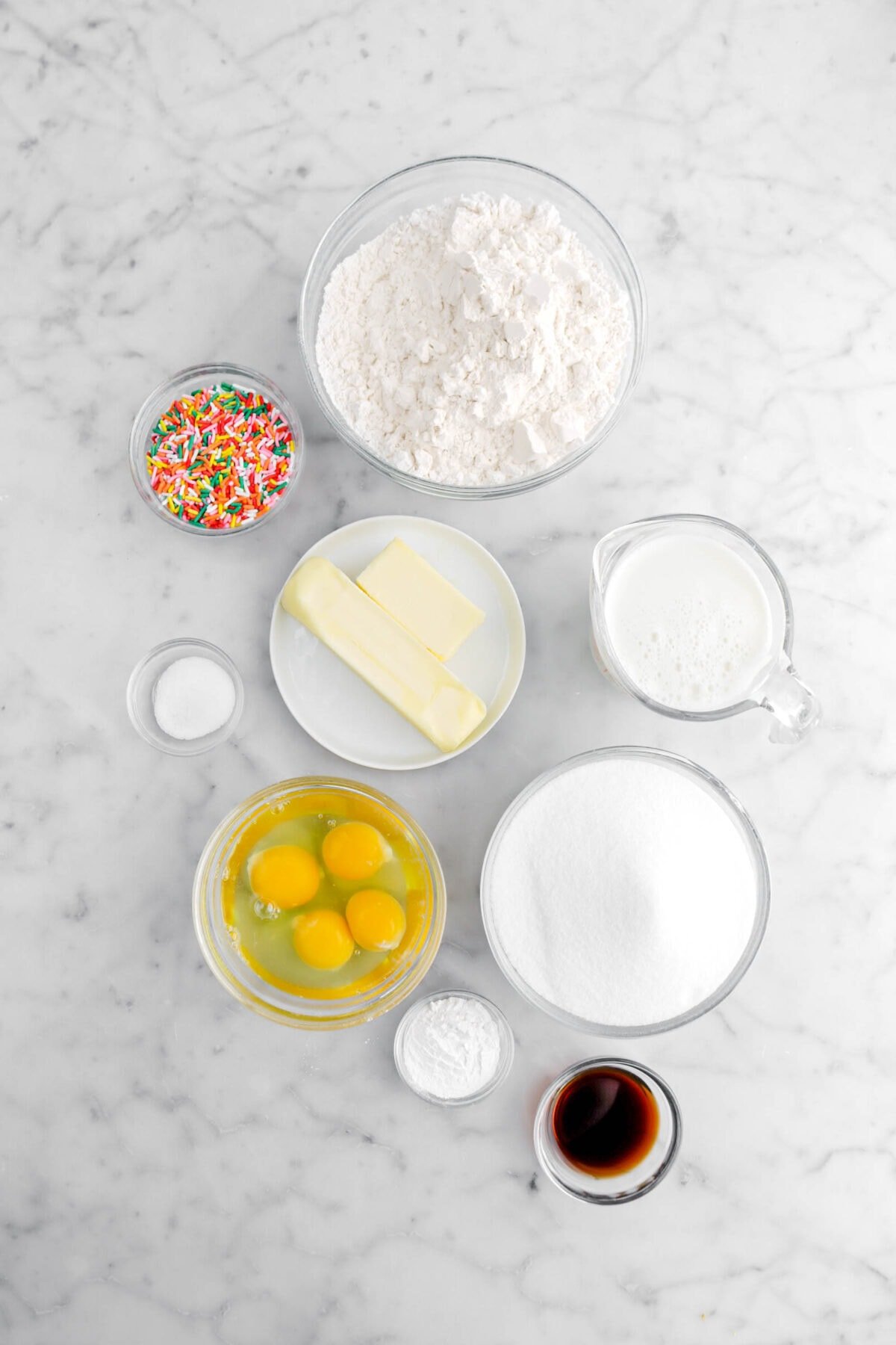 flour, rainbow sprinkles, butter, milk, salt, eggs, baking powder, sugar, and vanilla on marble surface.