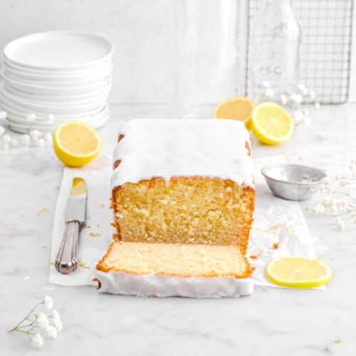 lemon loaf cake on parchment paper with slice laying in front with lemon halves behind, white flowers around, and stack of plates.