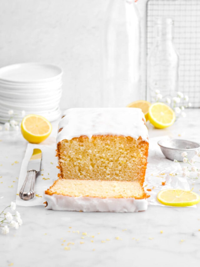 iced loaf cake on parchment paper with slice laying in front with a knife beside, white flowers around, and lemons behind on marble surface with stack of plates and empty glasses behind.