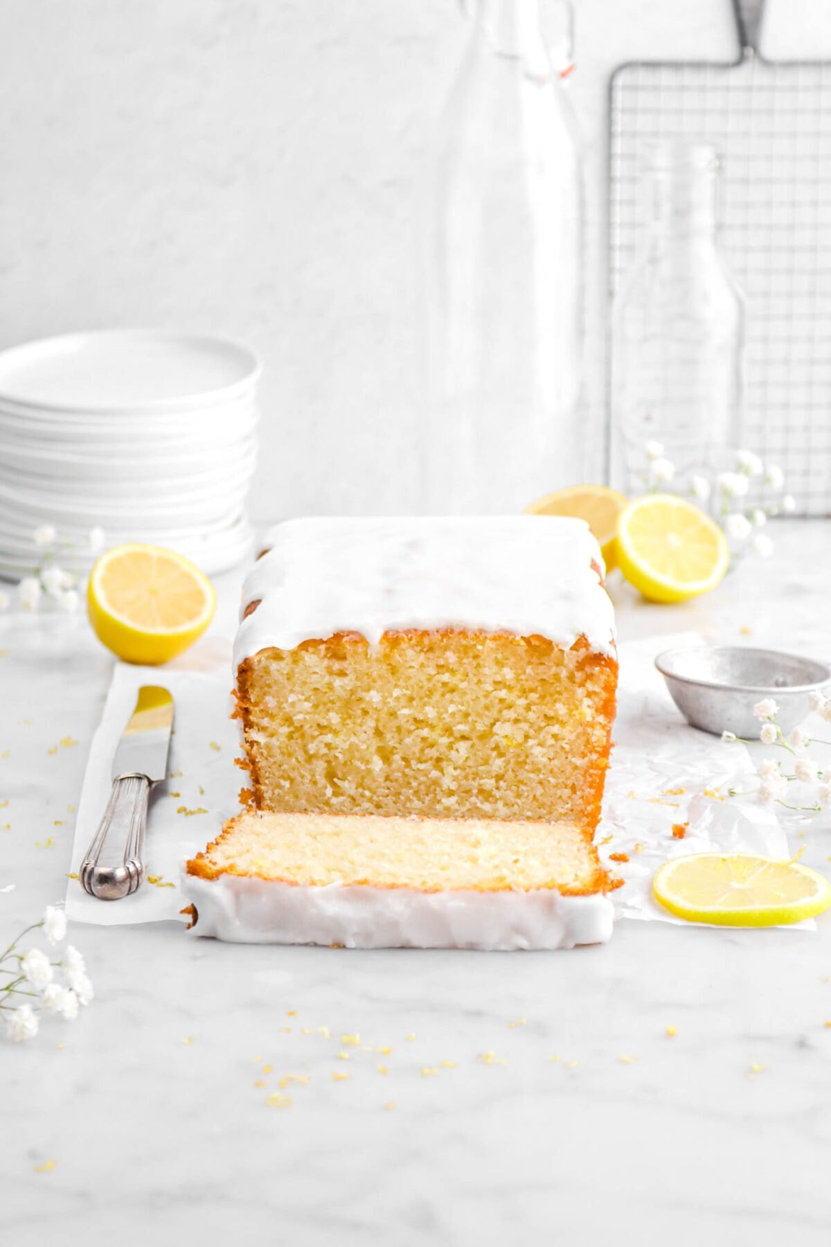 iced loaf cake on parchment paper with slice laying in front with a knife beside, white flowers around, and lemons behind on marble surface with stack of plates and empty glasses behind.