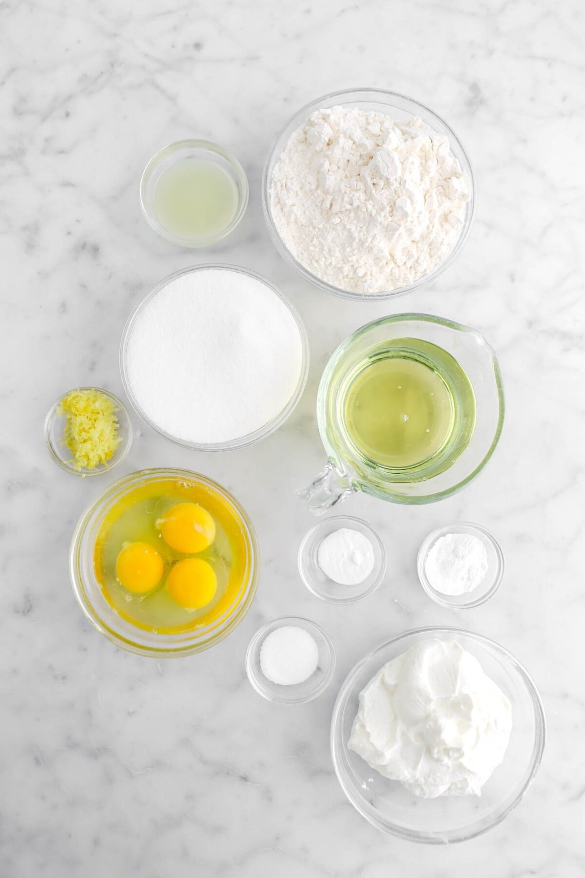 lemon juice, flour, sugar, lemon zest, oil, eggs, salt, baking soda, baking powder, and yogurt on marble surface.