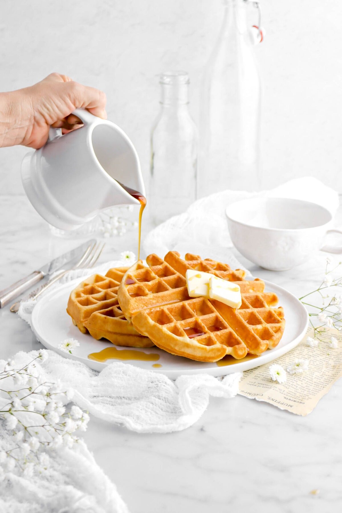 two waffles on large white plate with two pads of butter on one waffle with maple syrup being poured onto waffle, with white cheesecloth and white flowers around.