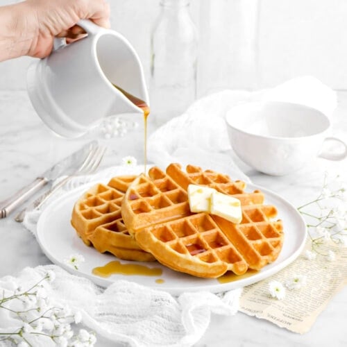 waffles on white plate on top of a white cheesecloth with maple syrup being poured onto one waffle, with white flowers, a mug, and two empty glasses behind.