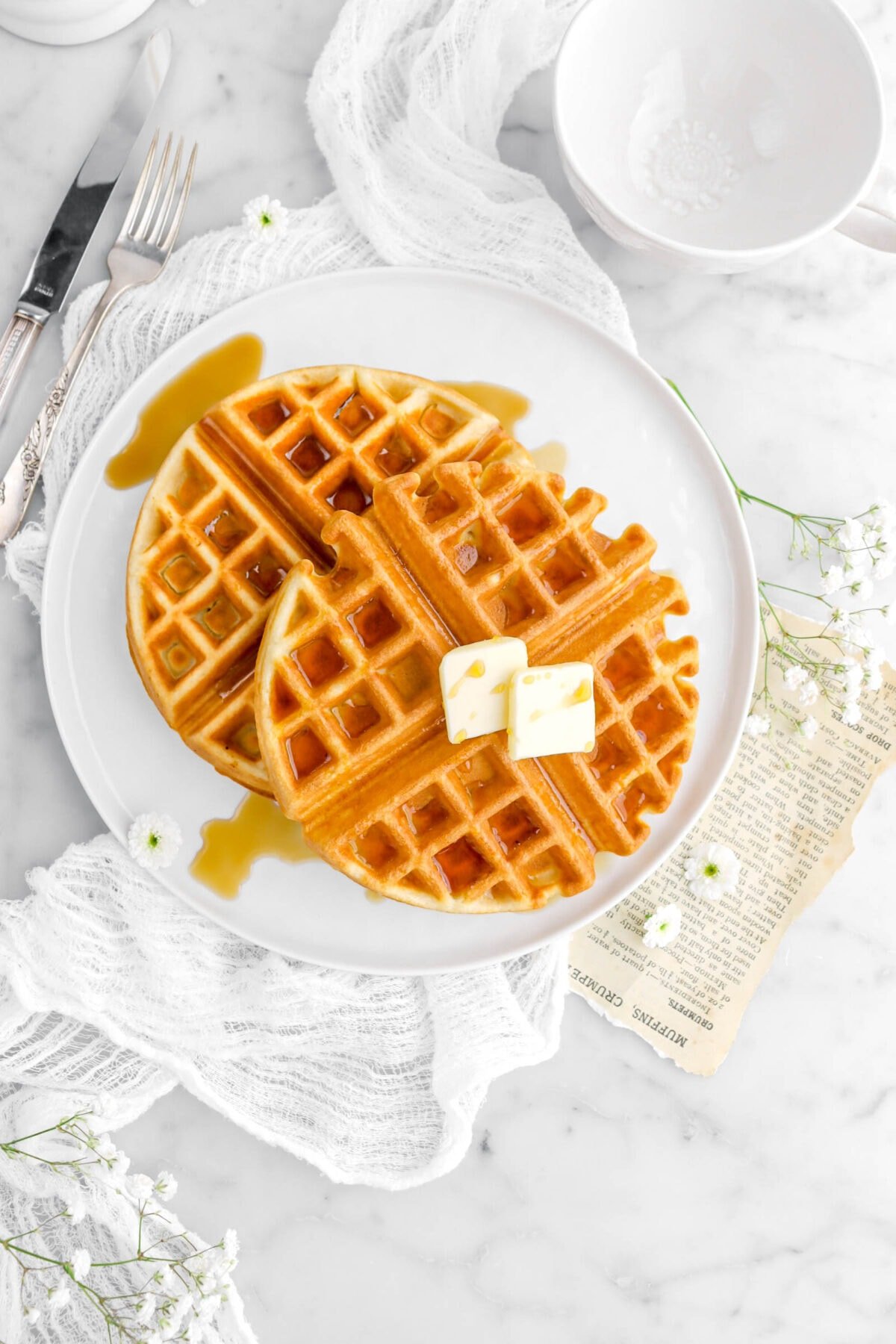 overhead shot of two waffles with maple syrup on both and two pats of butter on one on a white plate on top of a white cheesecloth with flowers around and an old book page.