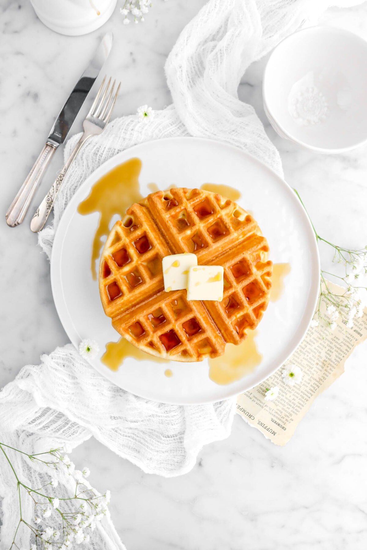 overhead shot of waffle with maple syrup and two pats of butter on top on a white plate, with fork and knife beside and white flowers around.