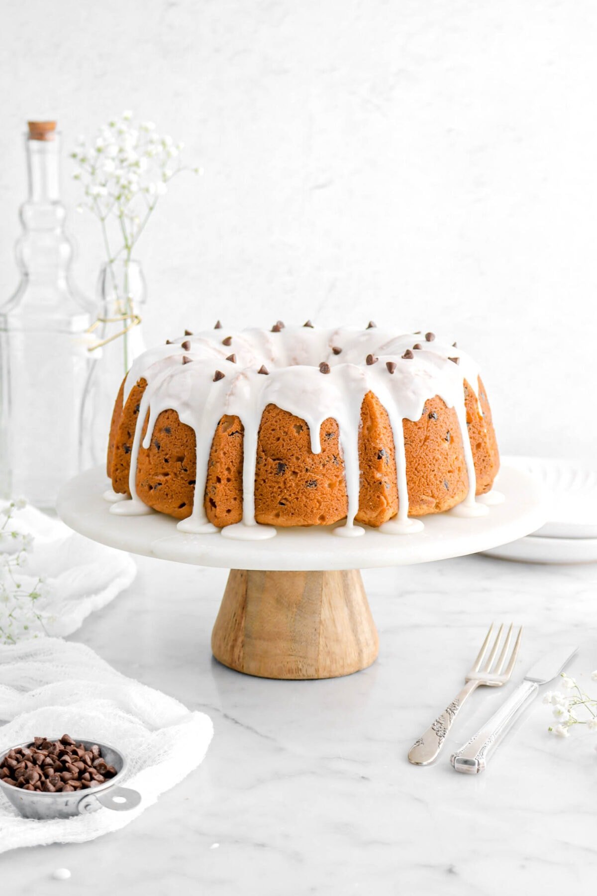 bundt cake on cake stand with icing and chocolate chips on top, a measuring cup of chocolate chips, a fork and knife, a white cheesecloth, and flowers beside.