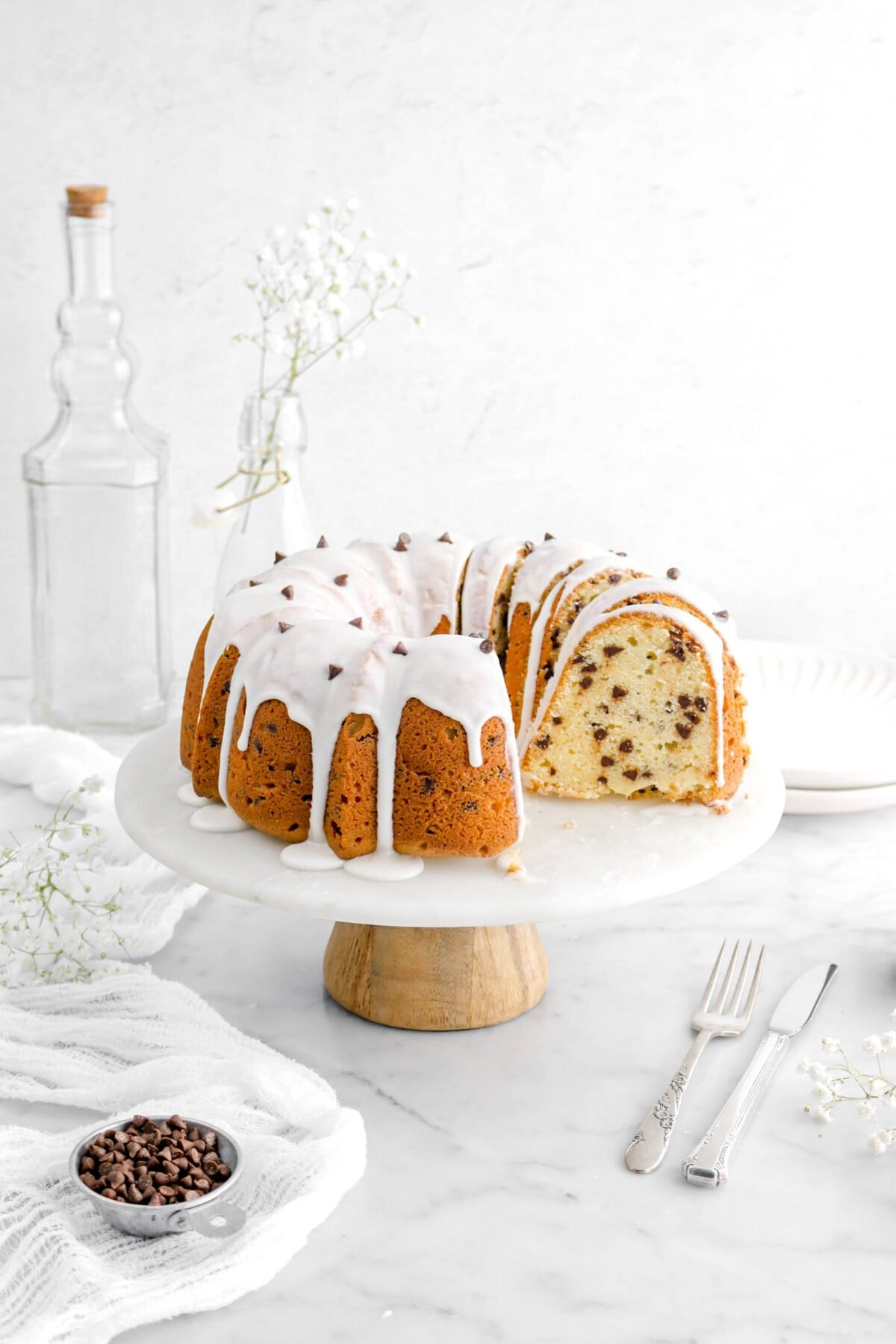 five slices of chocolate chip pound cake leaning against the rest of the cake on a cake stand, with stack of plates and empty glasses behind, a white cheesecloth beside, a measuring cup with mini chocolate chips, and a fork and knife beside on marble surface.