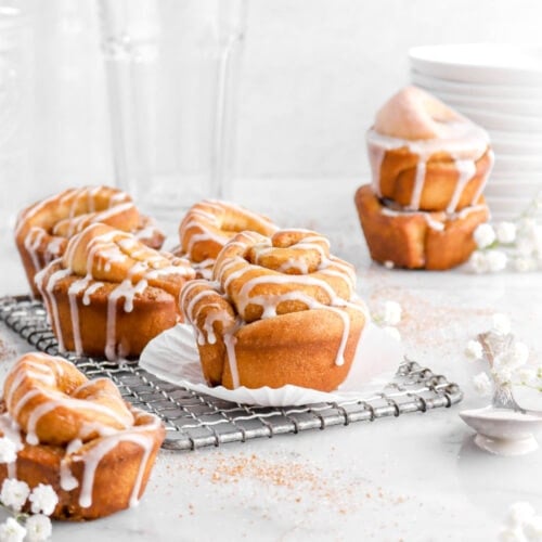 cinnamon rolls on wire cooling rack with white flowers and spoon beside, empty glasses and stack of plates behind.