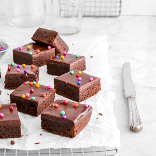brownies with ganache and rainbow chips on top, a knife beside, and empty glasses behind.