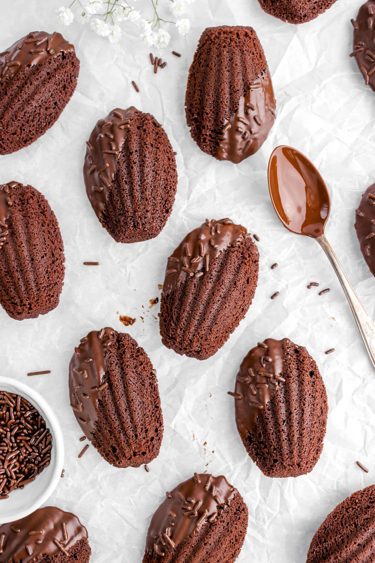 ten chocolate madeleines on parchment paper with spoon covered in chocolate beside and bowl of sprinkles beside.