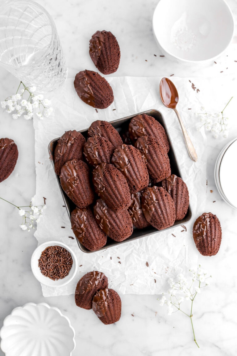 chocolate madeleines in square cake pan on parchment paper with more madeleines around, white flowers, chocolate sprinkles, an empty glass, a white mug, a stack of plates, and an empty scalloped owl around on marble surface.