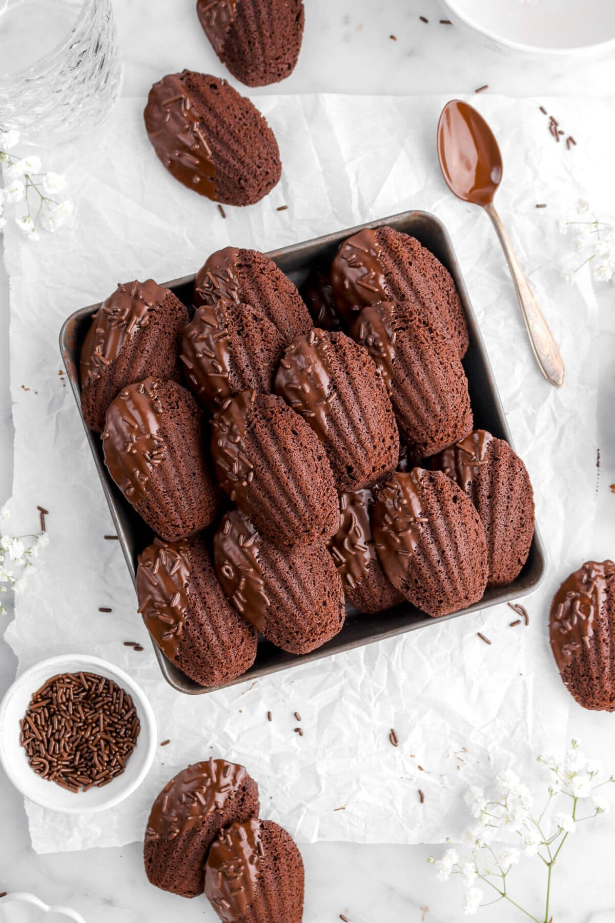 close up of chocolate madeleines in square pan on parchment paper with five more madeleines around, a bowl of sprinkles, white flowers, and a chocolate covered spoon.