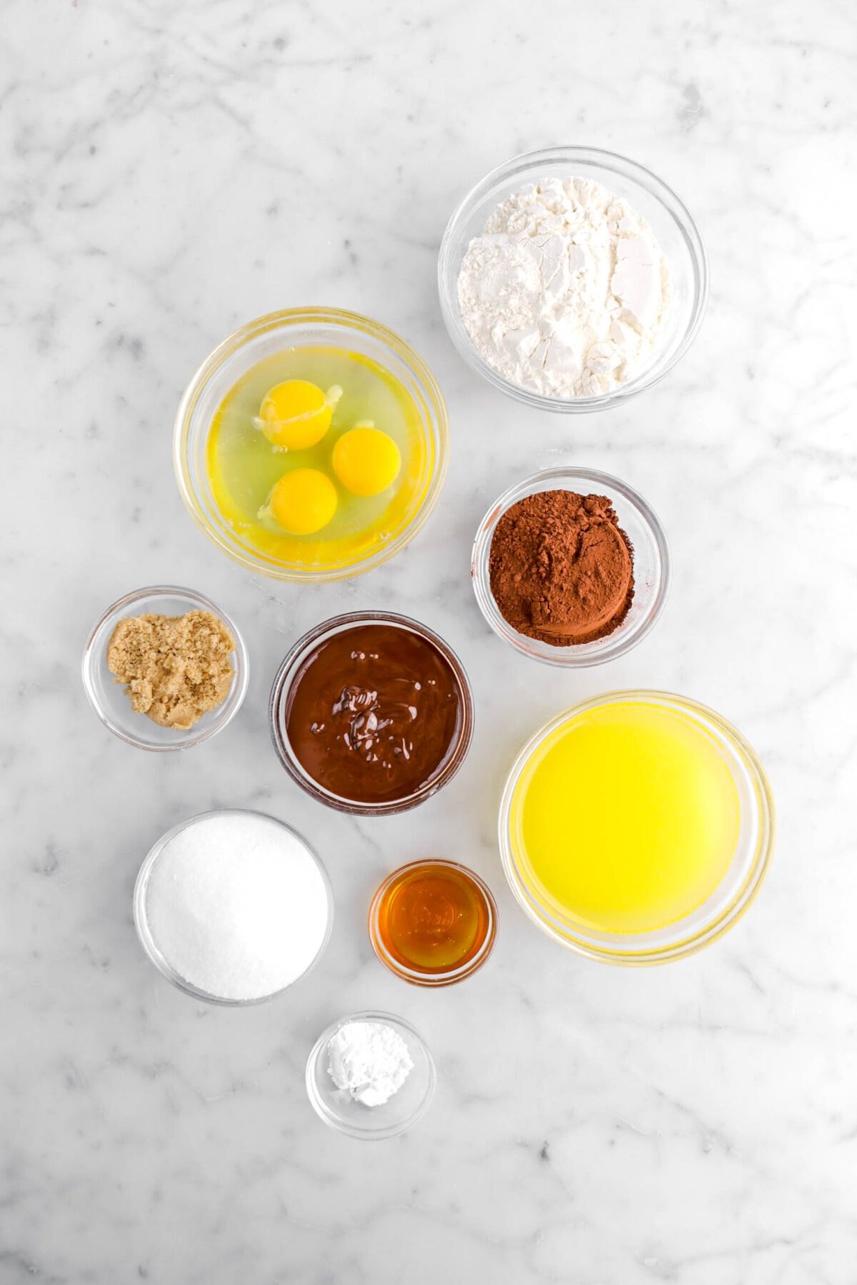 flour, eggs, cocoa powder, melted chocolate, brown sugar, granulated sugar, melted butter, honey, and baking powder in glass bowls on marble surface.