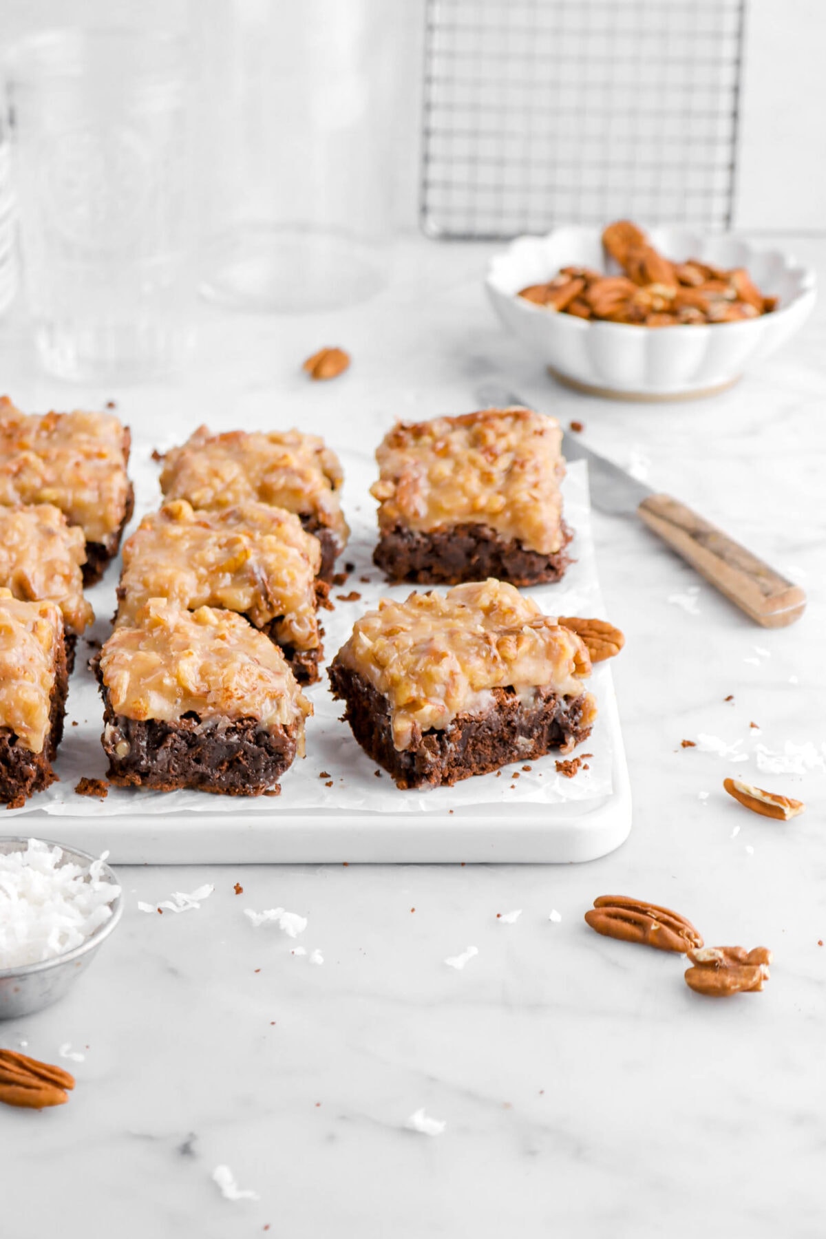 eight german chocolate brownies on parchment paper lined white tray with pecans and shredded coconut around on marble surface, with a wooden handled knife and bowl of pecans behind.