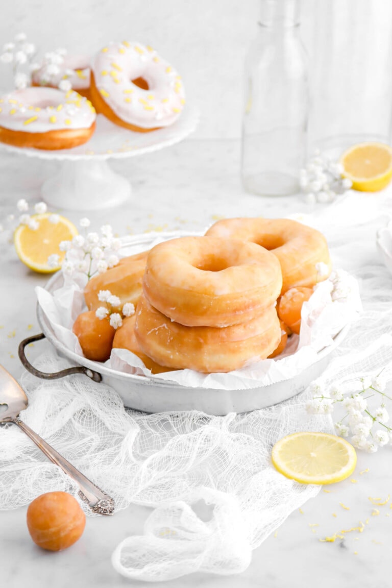 angled shot of lemon doughnuts in antique cake pan with parchment paper. Cheesecloth, white flowers, and lemon halves around on marble surface.