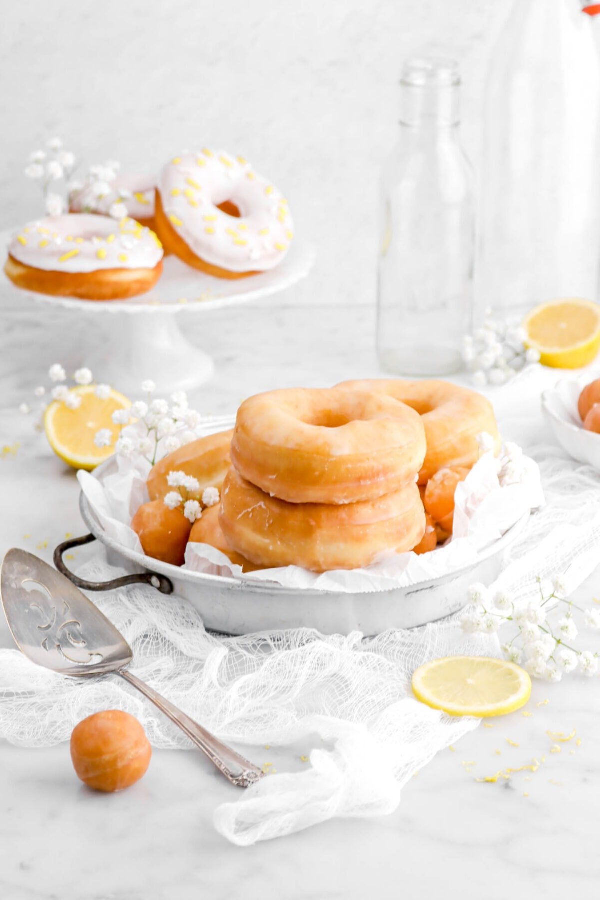 lemon doughnuts stacked in old cake pan on top of a white cheesecloth with cake knife beside and doughnut hole. Three dipped doughnuts on white cake stand behind with lemon halves and flowers.