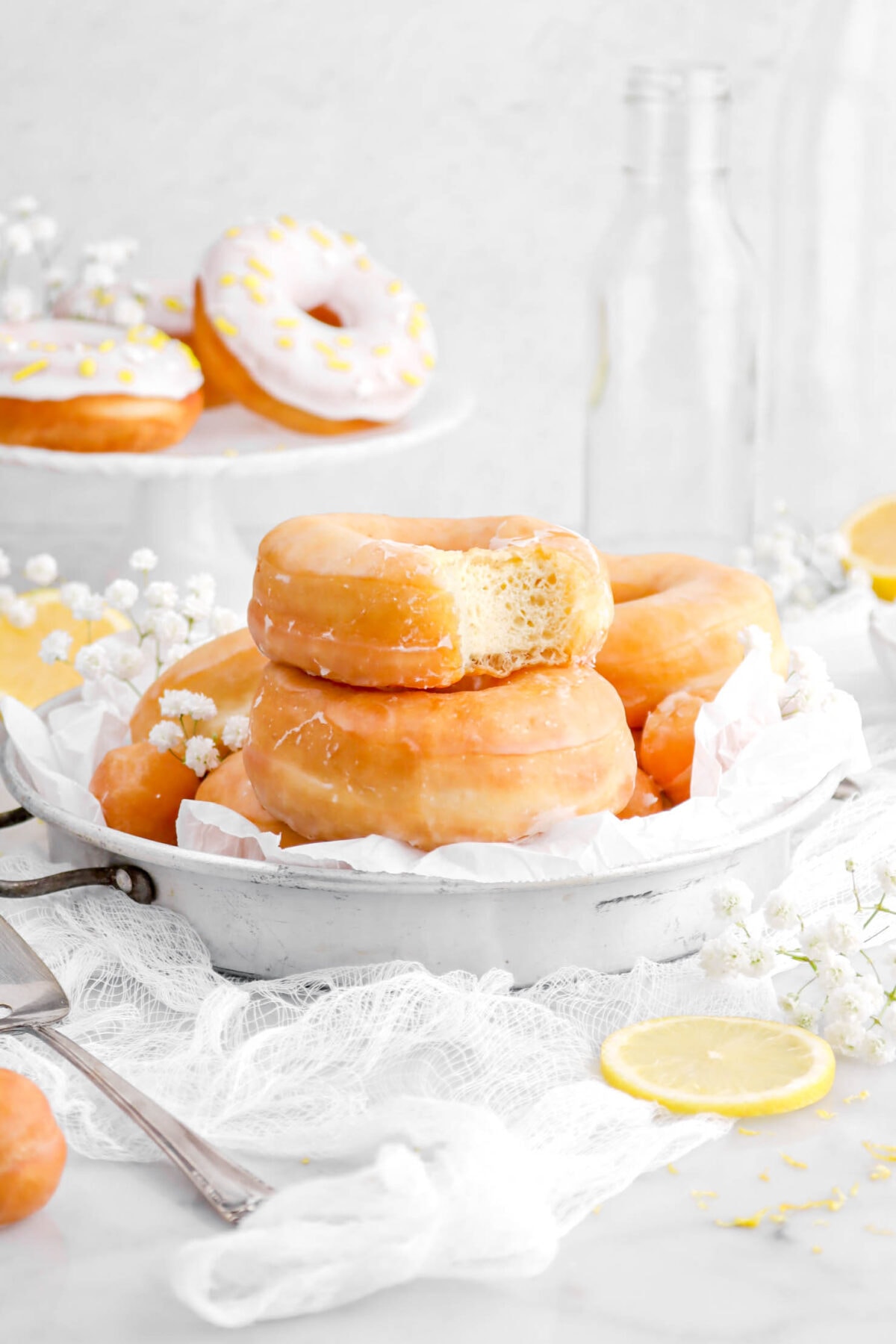 stacked lemon doughnuts in antique cake pan with top doughnut missing a bite, with white flowers and more doughnuts around.