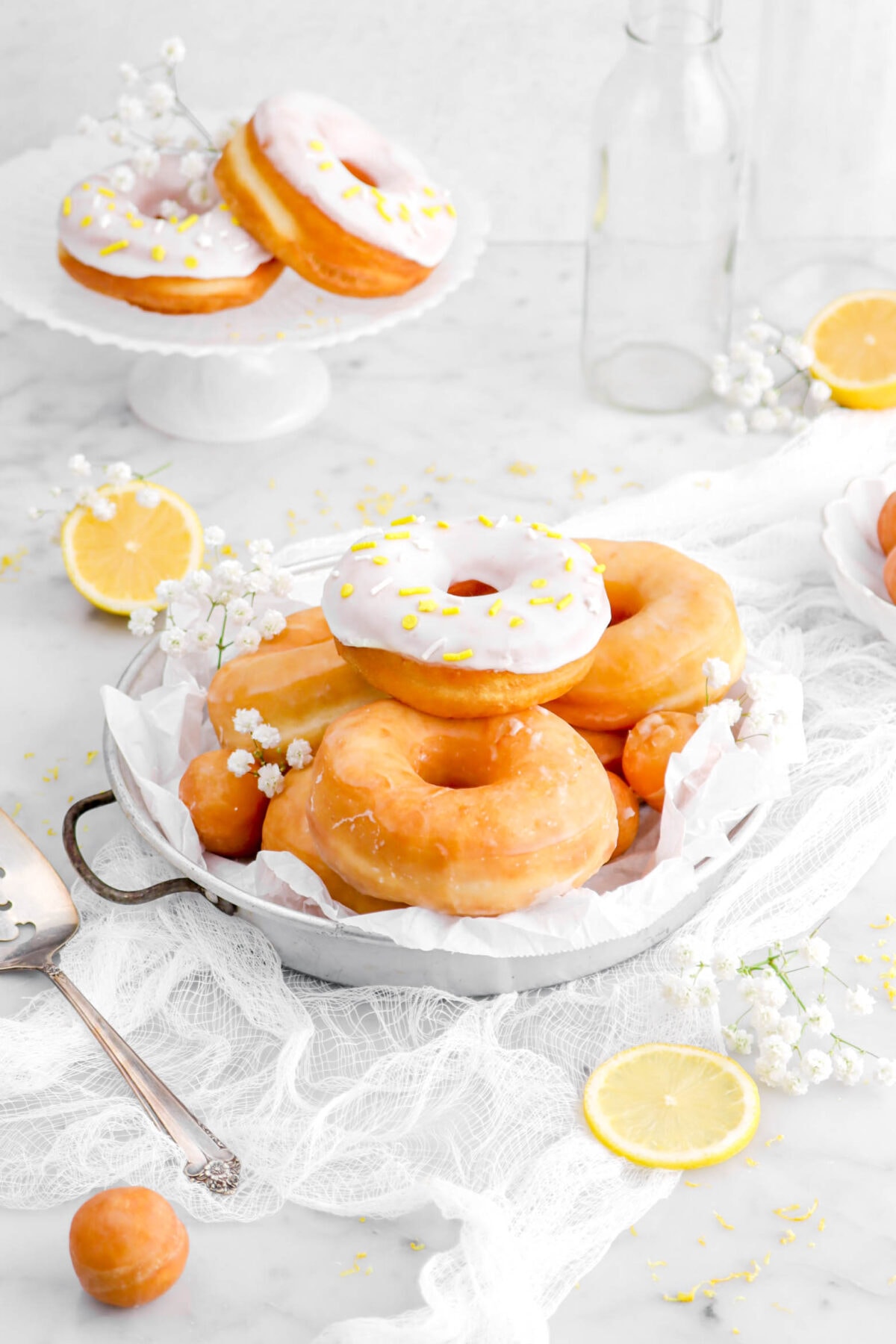 angled shot of doughnuts piled in cake pan on top of a white cheesecloth with lemon halves and white flowers around, and two doughnuts on cake plate behind.
