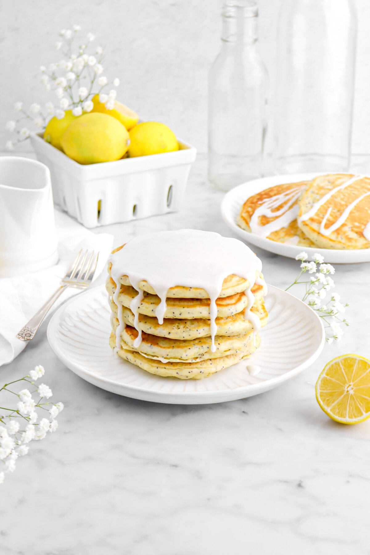 stack of lemon poppy seed pancakes on white plate with flowers around, a fork beside, a plate of pancakes behind, and berry basket full of lemons on marble surface.
