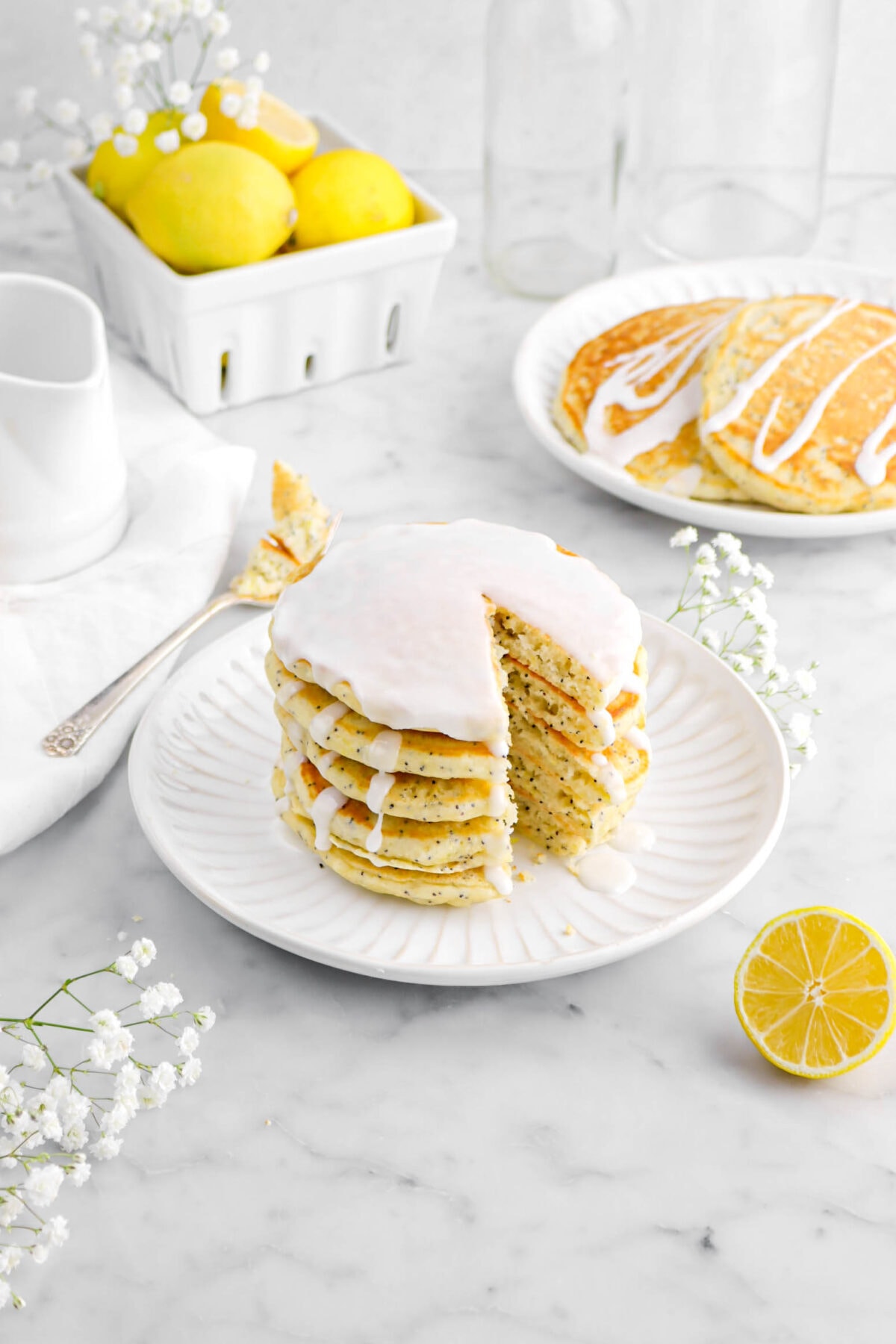 angled photo of sliced stacked pancakes with icing on top, a forkful of pancake beside, more pancakes behind on white plate, with flowers and lemons behind on marble surface.