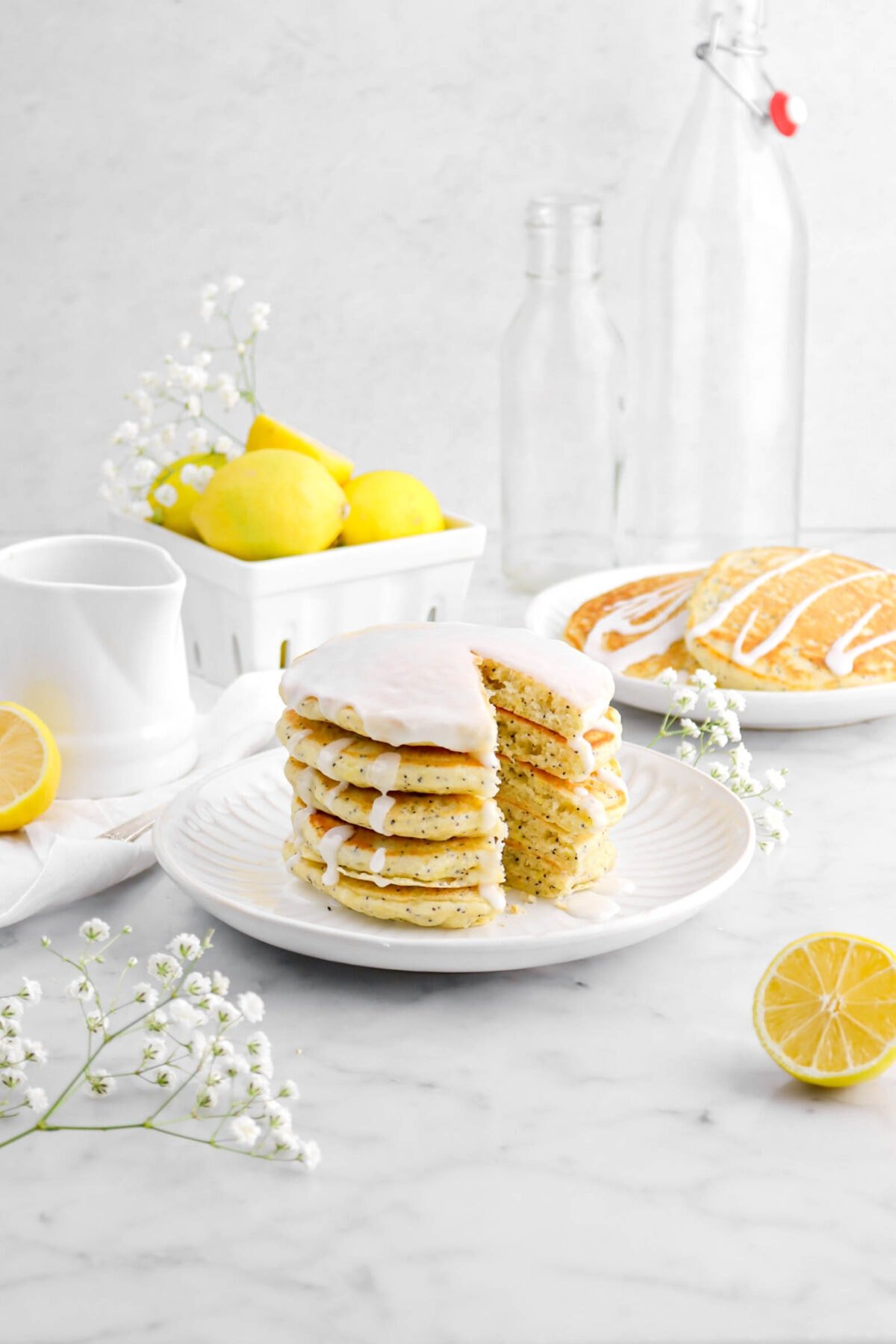 pulled back shot of stacked lemon poppy seed pancakes with icing dripping down the sides, lemons and white flowers around, and another plate of pancakes behind.
